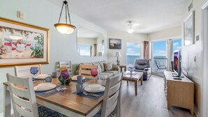 A dining area with a wooden table, chairs, and a sofa. There is a balcony with a railing and a view of the sky.