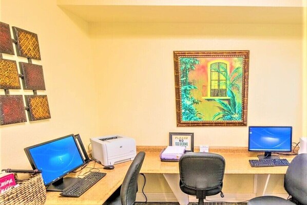 A workspace with two computer monitors, a printer, and a desk with a chair. There is a framed painting on the wall and a sign that says "PLEASE NO FOOD OR DRINKS".