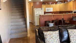 A living room with a black leather couch and patterned pillows, a staircase, a kitchen with wooden cabinets and a microwave, and a dining area with chairs. - 1A8 Views, Shared Pool & Hot Tub, Private Patio & Garage (Moab)