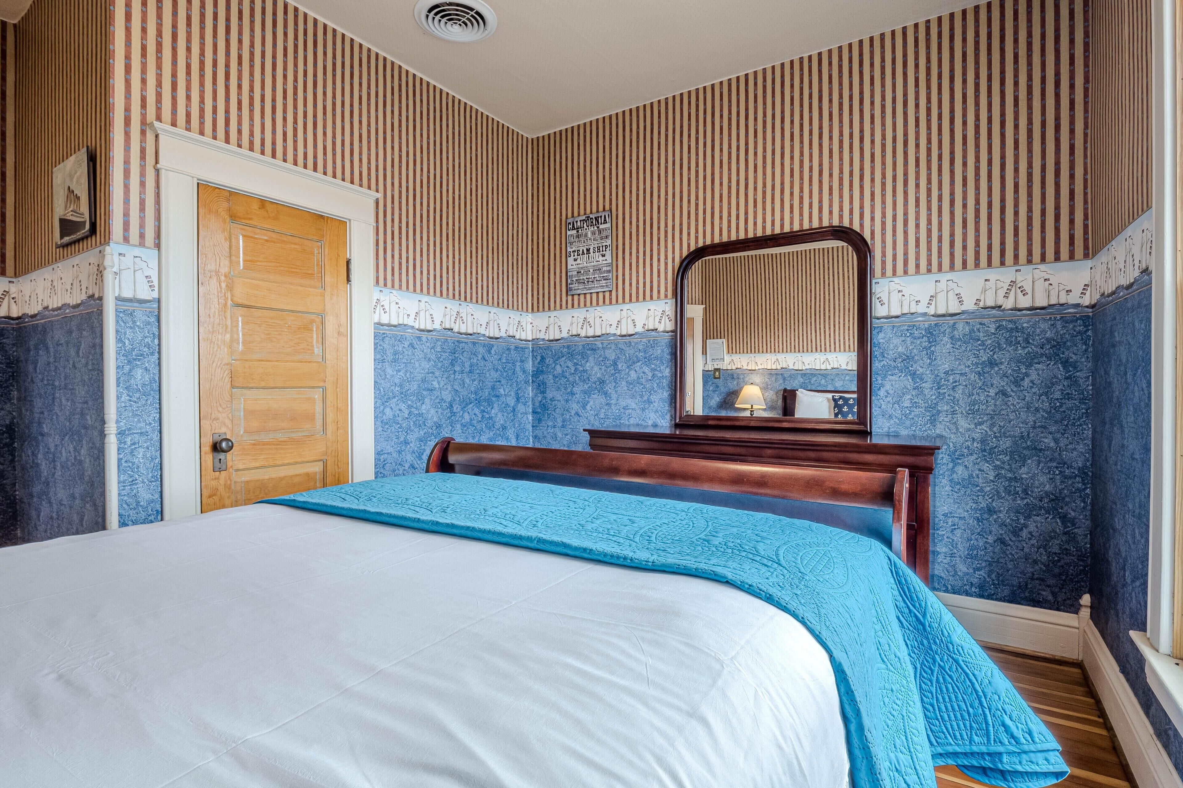 Une chambre avec une porte en bois, un lit recouvert d’une couverture bleue, une commode et un miroir au mur.