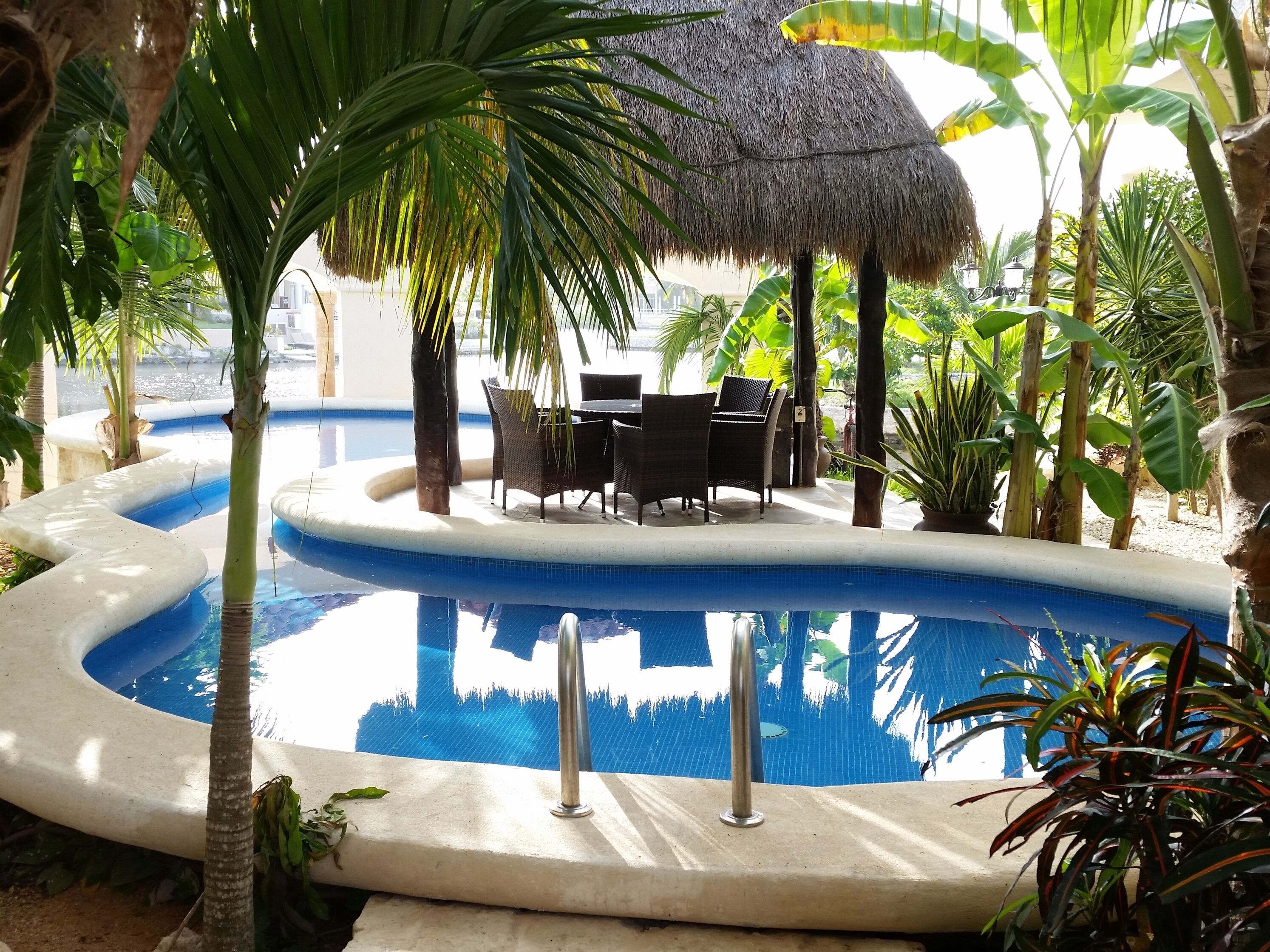 A pool area with a thatched roof, lounge chairs, and tropical plants.