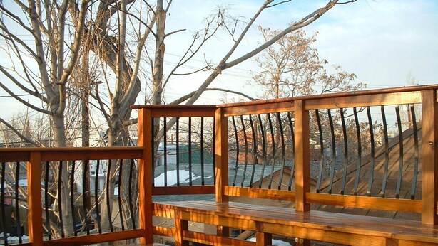 Una terraza de madera con un banco y barandilla, rodeada de árboles sin hojas y vista a un estacionamiento.
