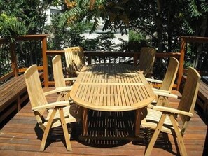 Ein runder Holztisch mit sechs Stühlen, umgeben von einer Holzterrasse mit Geländern, vor einer Kulisse aus üppigem Grün.