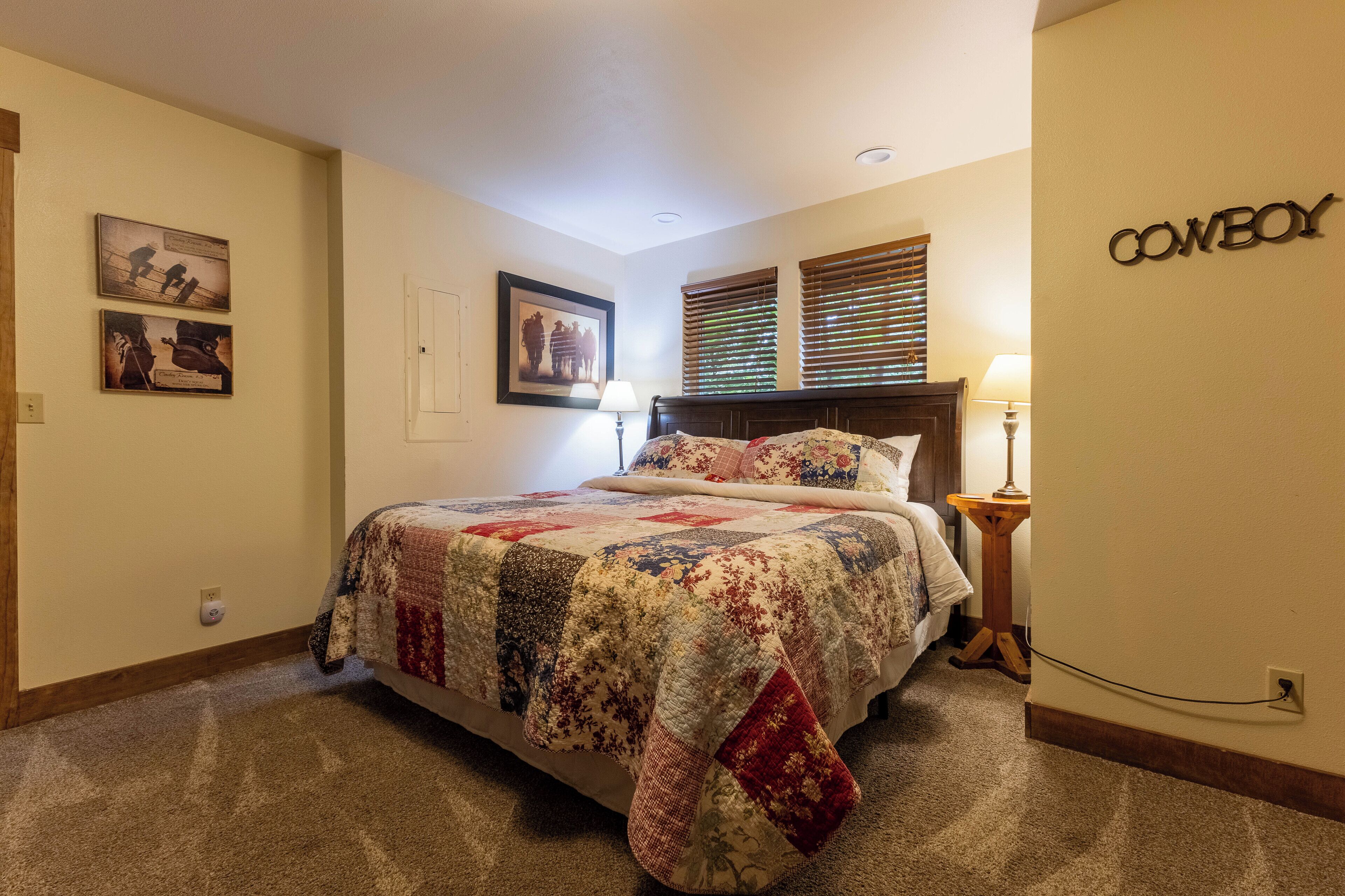 A bedroom with a large bed, a quilt, a nightstand with a lamp, and a wall-mounted sign that reads "COWBOY".