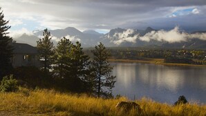 Vista de un lago con montañas al fondo, una casa y una zona boscosa.