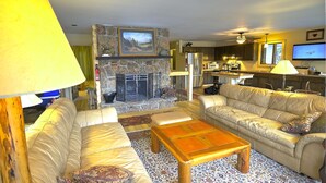 Una sala de estar con chimenea de piedra, sofás de cuero, una mesa de centro y una cocina al fondo.