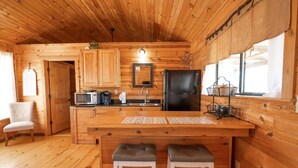 A wooden cabin interior with a kitchen area featuring a refrigerator, microwave, and wooden cabinets. There are two stools at the kitchen counter and a chair in the corner.