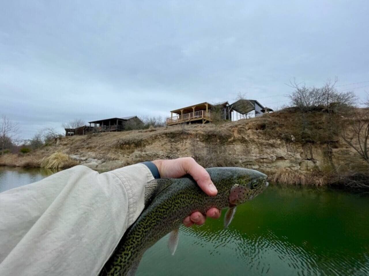 Une personne tenant un poisson près d’une étendue d’eau, avec une maison en arrière-plan.