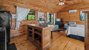 A cozy cabin interior with wooden walls, a ceiling fan, a kitchen area, a seating area with a TV, and a bed.