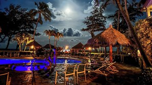 A nighttime resort pool area with thatched-roof structures, lounge chairs, and palm trees. - Direct Ocean Front, Top Floor Studio Paradise (Kapaa)