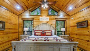 Dormitorio de una cabaña de madera con una cama, dos mesitas de noche, un ventilador de techo y una ventana con vista a árboles.