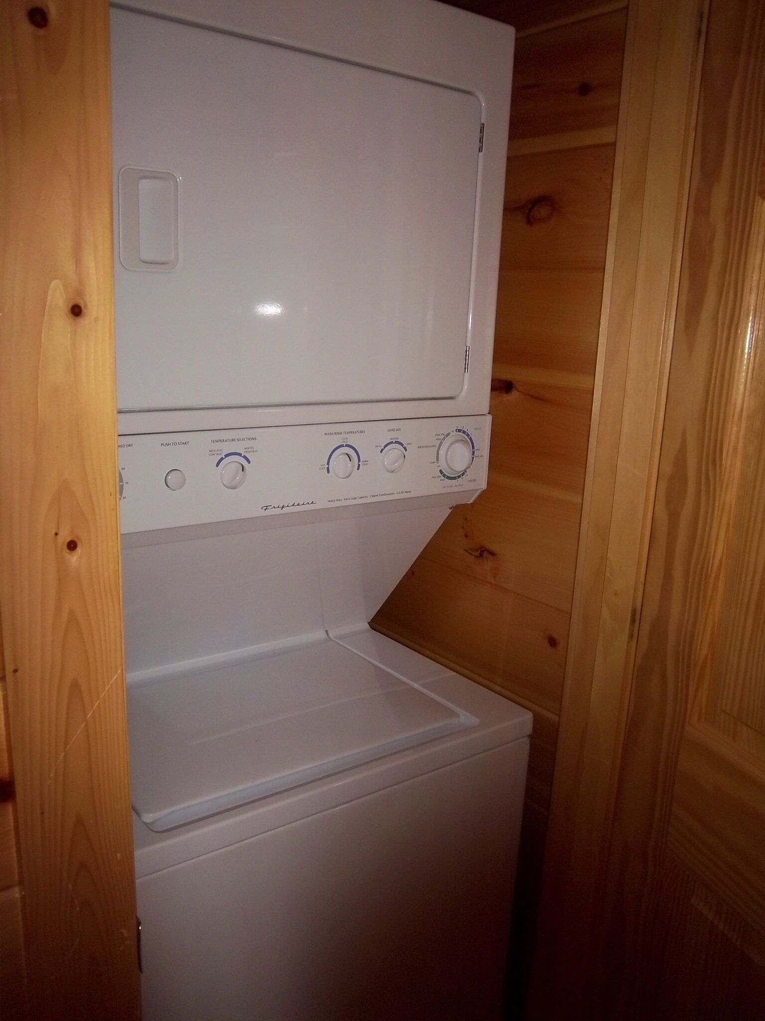 A compact, white washer and dryer unit with control knobs and a digital display, housed in a wooden-paneled room.