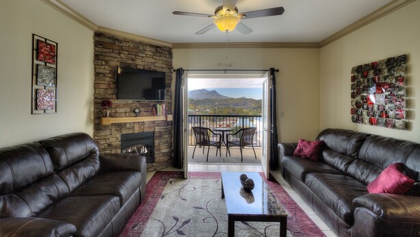 A living room with two leather sofas, a TV mounted on a stone wall, a fireplace, and a balcony with a mountain view. - John's Mtn Blessing  w/ 2 Pools, Hot tub, Arcade Games 7 BR & 6 BA.  Sleeps 20+ (Pigeon Forge)