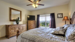 A bedroom with a bed, a dresser, a television, a ceiling fan, a window with curtains, and a framed picture on the wall. - John's Mtn Blessing  w/ 2 Pools, Hot tub, Arcade Games 7 BR & 6 BA.  Sleeps 20+ (Pigeon Forge)