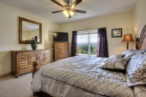 Quarto com cama, cômoda, televisão, ventilador de teto, janela com cortinas e luminária.