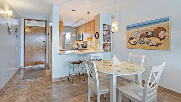 A dining area with a round table and white chairs, a kitchen in the background, and a beach-themed painting on the wall. - Oceanfront 2BR_2BA Kihei Maui–Stunning Lanai Sunsets, Beach Front, Turtles! (Kihei)