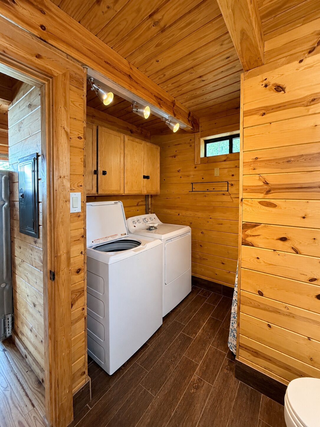 A compact wooden cabin interior with a washing machine, a refrigerator, and a small window.