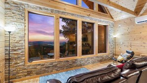 A cozy cabin with a stone wall, large windows, a leather sofa, and a view of the landscape. - Cliffside Escape | Epic Lake Views & Off-Grid Stay (Broken Bow)