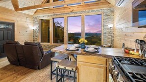 A cozy cabin interior with a stone fireplace, wooden dining table, and a large window overlooking a scenic landscape. - Cliffside Escape | Epic Lake Views & Off-Grid Stay (Broken Bow)
