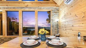 A wooden dining table with a view of a sunset through large windows, a vase with sunflowers, and a telephone on the table. - Cliffside Escape | Epic Lake Views & Off-Grid Stay (Broken Bow)