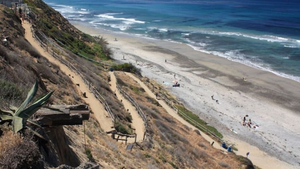 A coastal beach with a dirt path leading down to the shore, surrounded by dry vegetation and a cliff.
