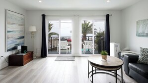 A modern living room with a sofa, a coffee table, and a TV. There is a balcony with a pool and outdoor furniture.