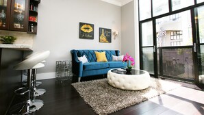 A modern living room with a blue sofa, a white ottoman, and a glass coffee table. There is a kitchen area with dark wood cabinets and a bar stool. Two wall art pieces are visible, one with a heart and the other with a building silhouette. - Luxury Condo in Santana Row - Silicon Valley - Levi's Stadium (San Jose)