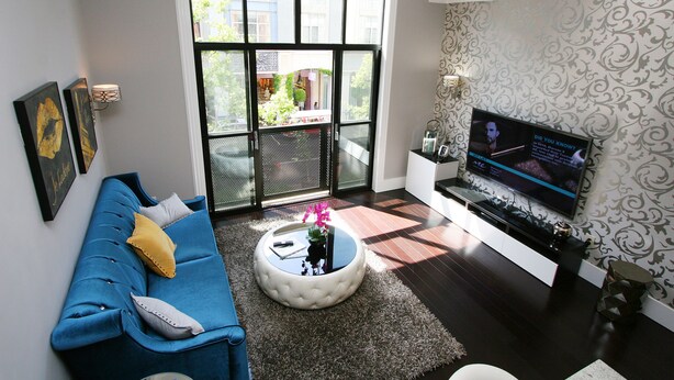 A living room with a blue sofa, a coffee table with a flower vase, a TV on the wall, and a sliding glass door leading to a balcony. - Luxury Condo in Santana Row - Silicon Valley - Levi's Stadium (San Jose)