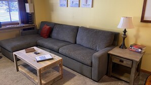 Una sala de estar acogedora con un sofá seccional gris, una mesa de centro de madera y una pequeña mesita de noche con una lámpara y libros.