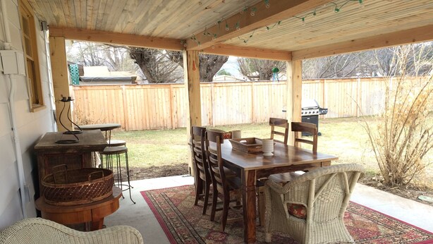 Ein überdachter Patio-Bereich mit Holztisch und -stühlen, Korbmöbeln und einem roten Teppich mit Muster.