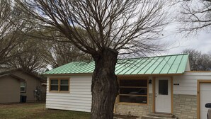 Ein eingeschossiges Haus mit einem grünen Metall-Dach, einem Steinfundament und einem großen Baum davor.