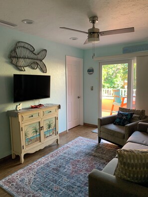 A living room with a flat-screen TV on a cabinet, a ceiling fan, a beach-themed cabinet, a patterned rug, and a sofa with decorative pillows.