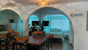 A dining area with a wooden table and chairs, a kitchen in the background, and a bedroom with a bed and a window. - Gulf side! No crossing the street to get to the beach! COQUINA BEACH CLUB 109 (Bradenton Beach)