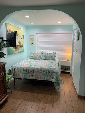 Une chambre à coucher comprenant un lit, un téléviseur fixé au mur, une fenêtre avec des stores, une table de chevet et une petite étagère.