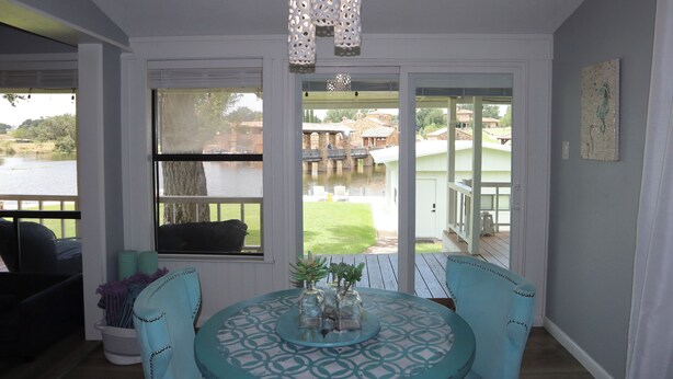 A room with a round table, chairs, a chandelier, and large windows with a view of the outdoors.