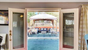 Puerta corredera de vidrio abierta que da a un patio exterior con zona de estar cubierta, piscina y alfombra roja.