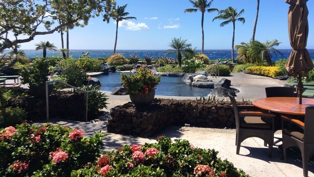 A resort pool area with a view of the ocean, surrounded by palm trees and tropical plants. - Relax by the Sea, in this awesome 2 bedroom Condo (Waikoloa)