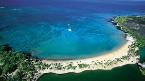 An aerial view of a beach with clear turquoise waters, a sandy shore lined with palm trees, and a small sailboat anchored near the center.