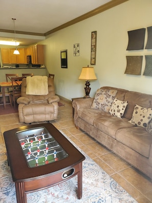 A living room with a foosball table, a sofa, and a coffee table. There is a kitchen in the background with wooden cabinets and a hanging light fixture.