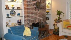 A living room with a brick fireplace, blue sofa, wooden coffee table, and various decorative items on shelves. - 'Beneath The Branches' Cottage Steps to Golf and Tennis. Pet-Friendly! (Murrells Inlet)