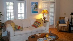 A living room with a white sofa, a coffee table, and a wicker chair. - 'Beneath The Branches' Cottage Steps to Golf and Tennis. Pet-Friendly! (Murrells Inlet)