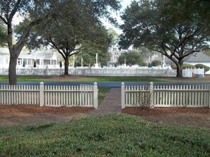 A park with white picket fences, trees, and a pathway leading to a building with a covered porch.