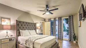 Un dormitorio con una cama grande, un ventilador de techo, una mesita de noche con lámpara y vistas al exterior a través de una puerta de vidrio.