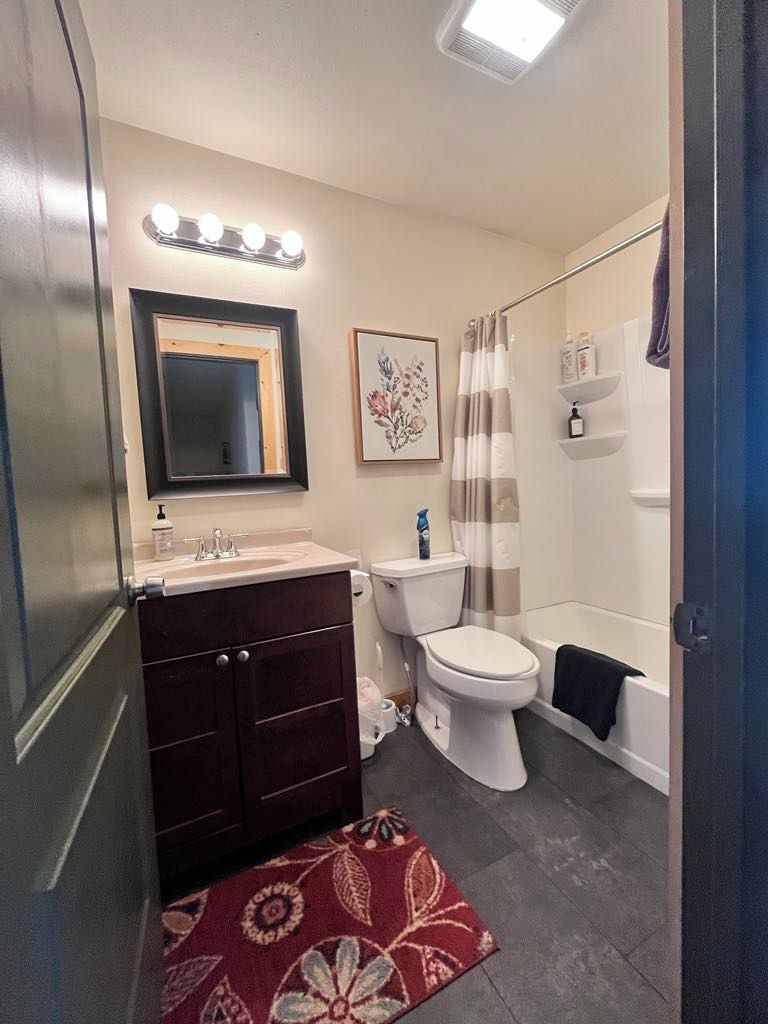 A bathroom with a dark wooden vanity, a white toilet, a shower with a curtain, and a small rug.