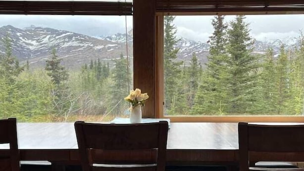 Un comedor con mesa y sillas, un florero con flores, y vistas a montañas nevadas y bosque.