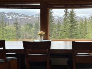En matplats med ett bord och stolar, en vas med blommor och utsikt över snöklädda berg och skog.