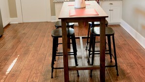Une cuisine avec une table en bois, des tabourets de bar, un luminaire suspendu et une fenêtre avec des stores.