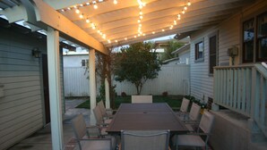 Une terrasse extérieure avec une table et des chaises en bois, des guirlandes lumineuses suspendues au-dessus et une clôture blanche en arrière-plan.