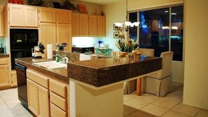 A kitchen with wooden cabinets, a granite countertop, a dishwasher, a sink, a dining area with a chair, and a window with a view of the outside. - Pond View Villa @ Desert Princess Country Club & Golf Resort (Cathedral City)