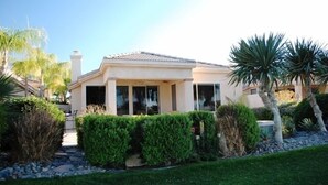 A single-story house with a tiled roof, a covered porch, and a well-maintained garden with various shrubs and palm trees. - Pond View Villa @ Desert Princess Country Club & Golf Resort (Cathedral City)